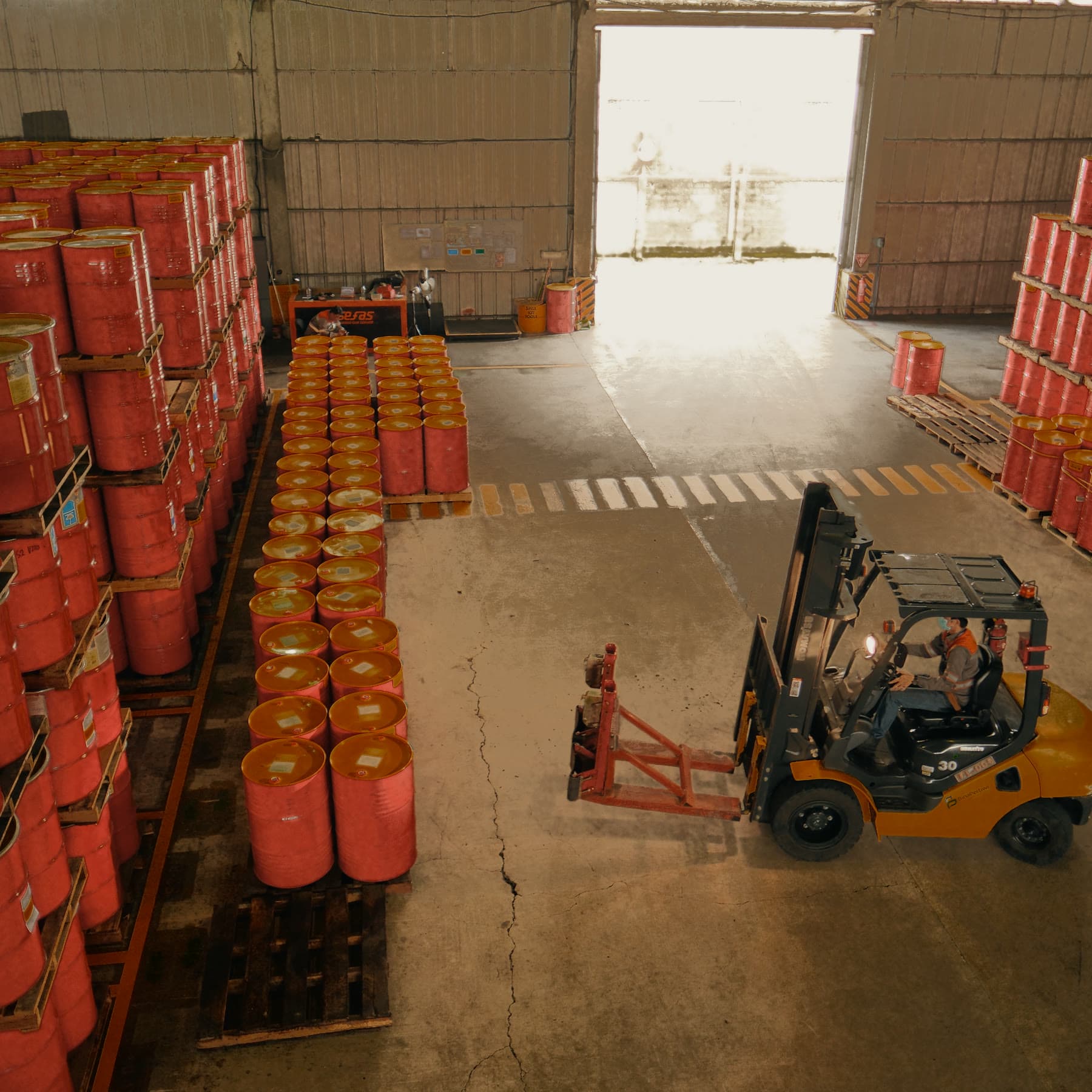 Forklift moving among stacked red-orange drums in a warehouse