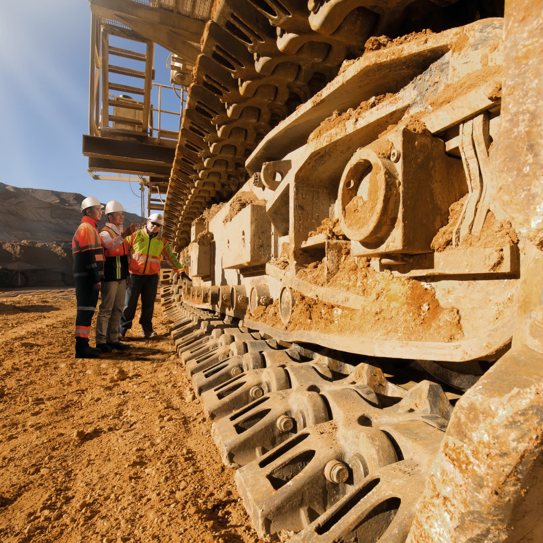 Three workers in safety gear beside the tracks of a large mining machine